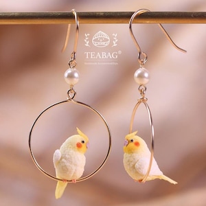 以下が含まれることがあります： 小さな黄色と白の鳥とオレンジ色の頬のパッチが特徴の、金色のフープイヤリング。各イヤリングにはパールのアクセントがあり、「TEABAG」というブランド名が見えます。