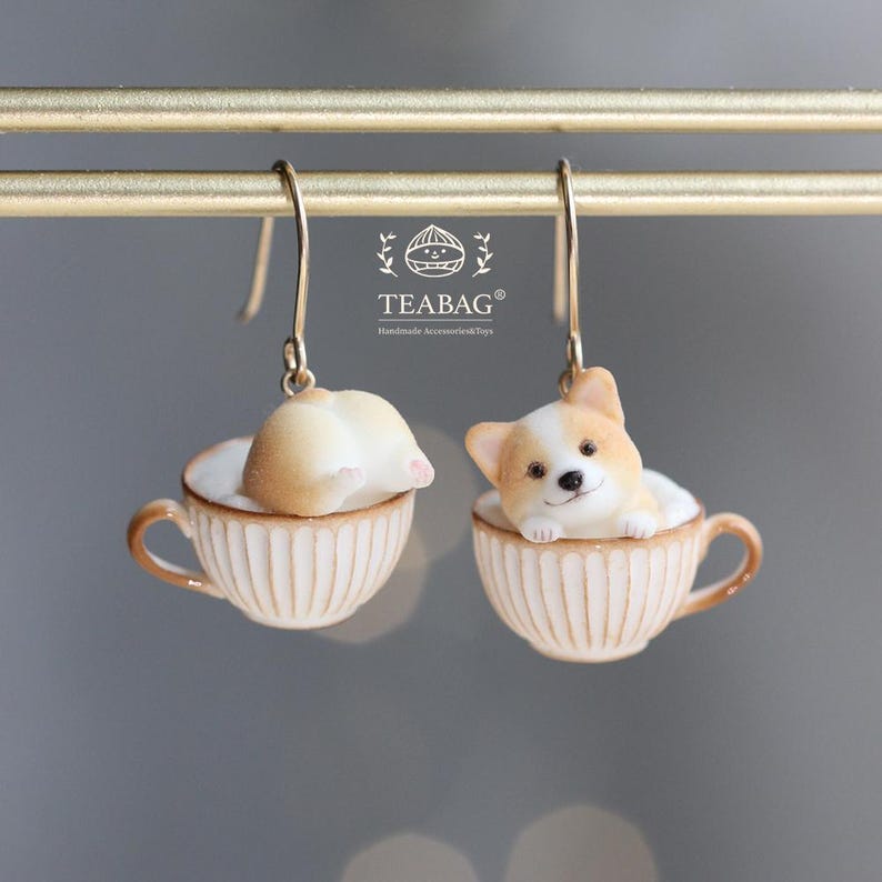 以下が含まれることがあります： ティーカップに入ったコーギー犬のフィギュアが特徴のぶら下がりピアス。 コーギーは白と茶色で、お尻を見せているものと顔を見せているものがあります。 ティーカップはクリーム色で、リブのディテールと金色のフックが付いています。