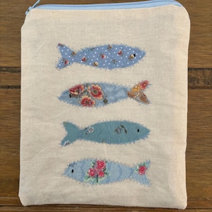 May include: A rectangular, beige fabric pouch with a light blue zip. The pouch features four appliquéd fish in shades of blue and floral patterns. The fish are stitched onto the fabric with visible thread.