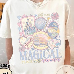 May include: Cream-coloured t-shirt with a pastel graphic design featuring a castle, Spaceship Earth, and other theme park icons. The text "MAGICAL PLACE ON EARTH" is displayed in blue and pink.