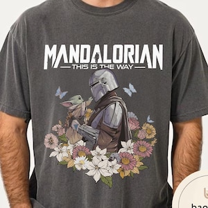 Puede incluir: Camiseta gris oscuro con un dise&ntilde;o de The Mandalorian y Grogu rodeados de flores y mariposas. El texto "MANDALORIAN - THIS IS THE WAY" est&aacute; impreso en blanco. El dise&ntilde;o incluye una ilustraci&oacute;n detallada de los personajes.