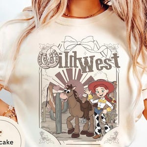 Pu&ograve; includere: T-shirt color crema con un design del West selvaggio. La grafica include una cowgirl dei cartoni animati, un cavallo e la scritta "Wild West" in stile vintage. Sono inclusi anche un ferro di cavallo e un cactus.