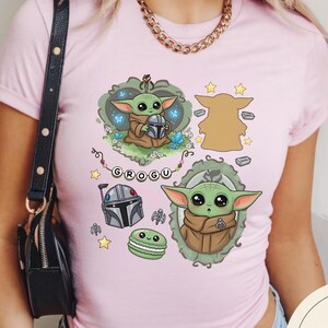 May include: A light pink t-shirt featuring a graphic print of the character Grogu in various poses, a Mandalorian helmet, a green macaron, and a string of beads spelling GROGU. The design includes decorative frames, small stars, and blue butterflies.