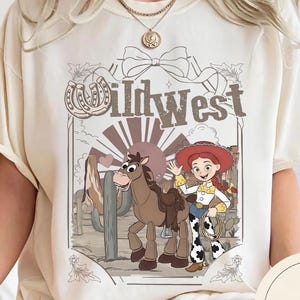Pu&ograve; includere: T-shirt color crema con un design del West selvaggio. La grafica include un cartone animato di una cowgirl con un lazo, un cavallo e la scritta "Wildwest". Il design ha uno stile vintage, a cartoni animati, con uno sfondo di paesaggio desertico.