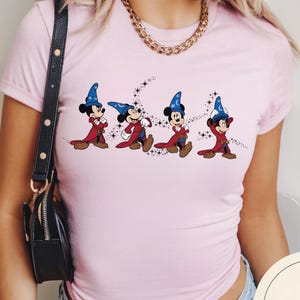 May include: A light pink t-shirt featuring a graphic of Mickey Mouse in a sorcerer's outfit. The cartoon characters are depicted in a row, wearing blue wizard hats and red robes, with a starry design.