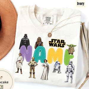 May include: Ivory-colored t-shirt featuring Star Wars characters and the text "NAME" in colorful letters. The shirt includes images of Darth Vader, Chewbacca, Yoda, and other characters from the Star Wars franchise. A gold necklace is draped on the shirt.