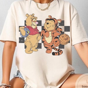 May include: Cream-colored t-shirt featuring a vintage-style illustration of Winnie the Pooh and Tigger dressed in Halloween costumes. Pooh wears a red shirt and carries a blue bag, while Tigger holds a carved pumpkin. The background has a black and white checkered pattern.