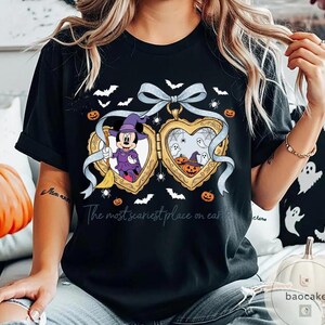 May include: Black t-shirt with a Halloween-themed heart-shaped locket design. The locket depicts Minnie Mouse in a witch costume and pumpkins. The text "The most scariest place on earth" is printed below the locket.