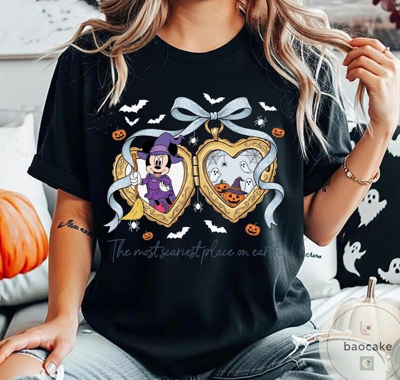 May include: Black t-shirt with a Halloween-themed heart-shaped locket design. The locket depicts Minnie Mouse in a witch costume and pumpkins. The text "The most scariest place on earth" is printed below the locket.