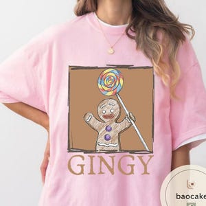 May include: Pink t-shirt featuring a gingerbread man holding a lollipop. The gingerbread man is brown with purple buttons and a smiling face. The word "GINGY" is printed in brown below the image.