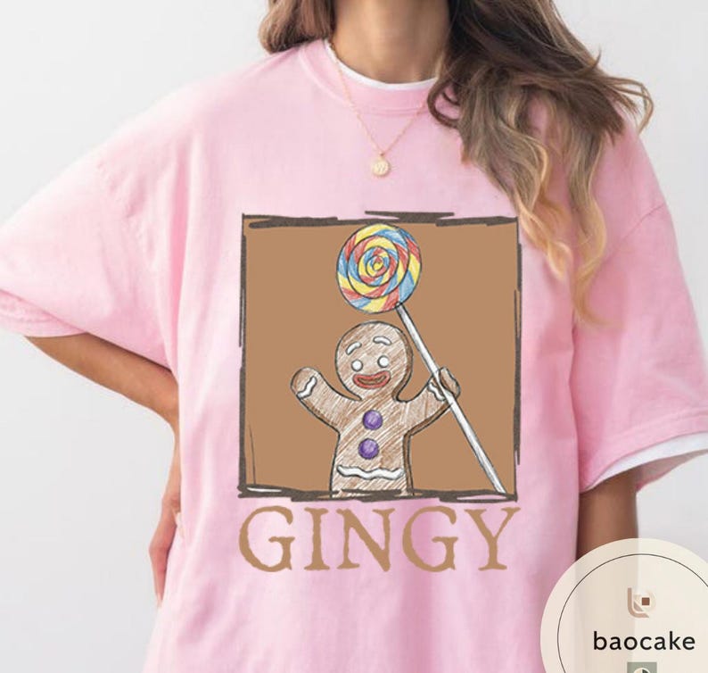 May include: Pink t-shirt featuring a gingerbread man holding a lollipop. The gingerbread man is brown with purple buttons and a smiling face. The word "GINGY" is printed in brown below the image.