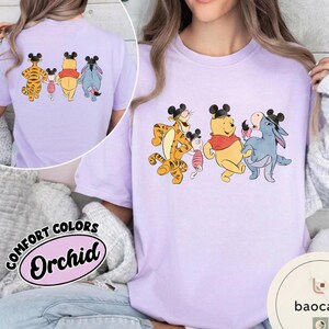 May include: A light purple t-shirt featuring a cartoon graphic of Winnie the Pooh characters wearing mouse ears. The characters include Tigger, Piglet, Winnie the Pooh, and Eeyore. The shirt also has the words "COMFORT COLORS" and "Orchid" printed on it.