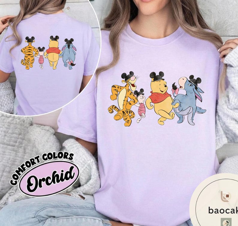 May include: A light purple t-shirt featuring a cartoon graphic of Winnie the Pooh characters wearing mouse ears. The characters include Tigger, Piglet, Winnie the Pooh, and Eeyore. The shirt also has the words "COMFORT COLORS" and "Orchid" printed on it.