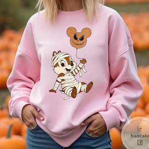 May include: A light pink sweatshirt featuring a cartoon character wrapped in bandages, holding a brown balloon with a jack-o'-lantern face. The design is a Halloween theme. The background is a pumpkin patch.