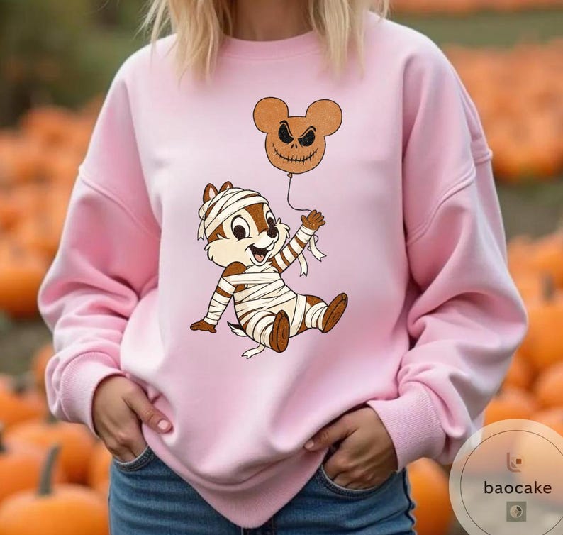 May include: A light pink sweatshirt featuring a cartoon character wrapped in bandages, holding a brown balloon with a jack-o'-lantern face. The design is a Halloween theme. The background is a pumpkin patch.