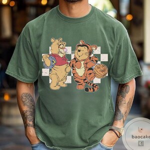 May include: Green t-shirt featuring a cartoon illustration of Winnie the Pooh and Tigger. Winnie the Pooh is wearing a red shirt and holding a blue backpack. Tigger is holding a pumpkin. The background is a white and black checkered pattern.