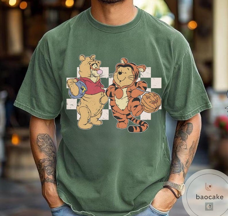 May include: Green t-shirt featuring a cartoon illustration of Winnie the Pooh and Tigger. Winnie the Pooh is wearing a red shirt and holding a blue backpack. Tigger is holding a pumpkin. The background is a white and black checkered pattern.