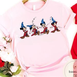 May include: Light pink t-shirt featuring a graphic of Mickey Mouse in a sorcerer's outfit. The cartoon character is depicted in a series of poses, with a blue hat, red robe, and brown shoes. Black stars trail behind each figure, creating a magical effect.