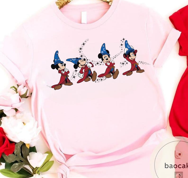 May include: Light pink t-shirt featuring a graphic of Mickey Mouse in a sorcerer's outfit. The cartoon character is depicted in a series of poses, with a blue hat, red robe, and brown shoes. Black stars trail behind each figure, creating a magical effect.