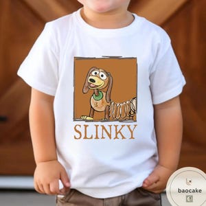 K&ouml;nnte beinhalten: Ein wei&szlig;es Kurzarm-T-Shirt mit einer quadratischen Grafik eines braunen Zeichentrickhundes mit federartigem K&ouml;rper. Der Hund tr&auml;gt ein gr&uuml;nes Halsband und befindet sich in einem braunen Rahmen. Das Wort SLINKY steht in braunen Gro&szlig;buchstaben darunter.