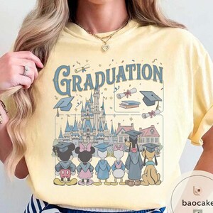 May include: Yellow t-shirt with a graphic featuring the word "Graduation" in blue script, a castle, and cartoon characters in graduation attire. The design includes Mickey and Minnie Mouse, Donald and Daisy Duck, and Pluto.