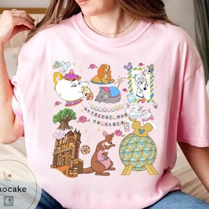 May include: Pink t-shirt featuring a colorful graphic with cartoon characters, including a teapot, dogs, cats, and a kangaroo. The text "Happiest Mom on Earth" is also included in the design. The shirt is a casual, everyday wear item.