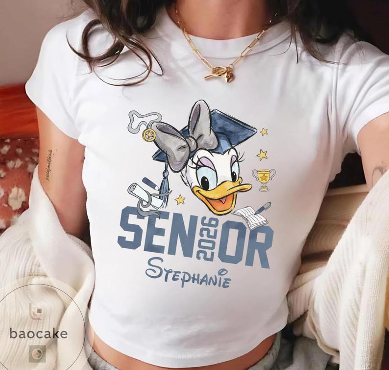 May include: White t-shirt featuring a cartoon Daisy Duck wearing a graduation cap and bow. The shirt has the text "SENIOR 2026 Stephanie" in blue. Additional graphics include stars, a trophy, and a diploma.
