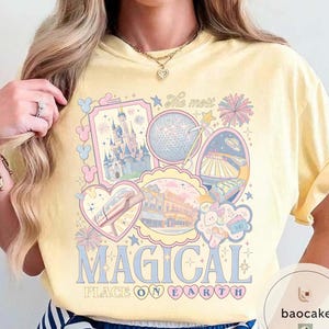 May include: A pale yellow t-shirt with a colourful graphic design. The design features the text "The most MAGICAL PLACE ON EARTH" and illustrations of a castle, a sphere, and other theme park elements in pastel colours.