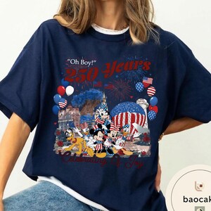 Puede incluir: Camiseta azul marino con un dise&ntilde;o de personajes de Disney, fuegos artificiales y globos. La camiseta muestra el texto "Oh Boy! 250 Years Celebrating of Joy". El dise&ntilde;o incorpora banderas americanas y colores patri&oacute;ticos.