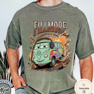 May include: Sage green t-shirt with a cartoon illustration of a green VW bus. The text "FILLMORE" is above the bus, which has a rainbow peace sign and the number 95. The design includes lightning bolts and tools.