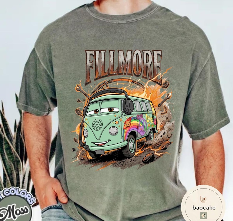 May include: Sage green t-shirt with a cartoon illustration of a green VW bus. The text "FILLMORE" is above the bus, which has a rainbow peace sign and the number 95. The design includes lightning bolts and tools.