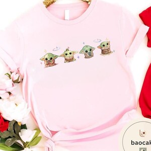 May include: A light pink t-shirt featuring a graphic of four cartoon characters. The characters are green with large ears and are in different poses. The shirt has short sleeves and a round neckline. The shirt is made of a soft material.