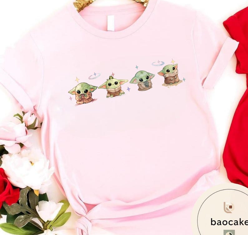 May include: A light pink t-shirt featuring a graphic of four cartoon characters. The characters are green with large ears and are in different poses. The shirt has short sleeves and a round neckline. The shirt is made of a soft material.