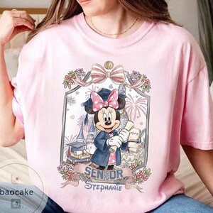 May include: Pink t-shirt featuring a Minnie Mouse graduation design. Minnie Mouse is wearing a graduation cap and gown, holding a diploma. The design includes the text "Senior Stephanie" and floral accents, with a castle and fireworks in the background.
