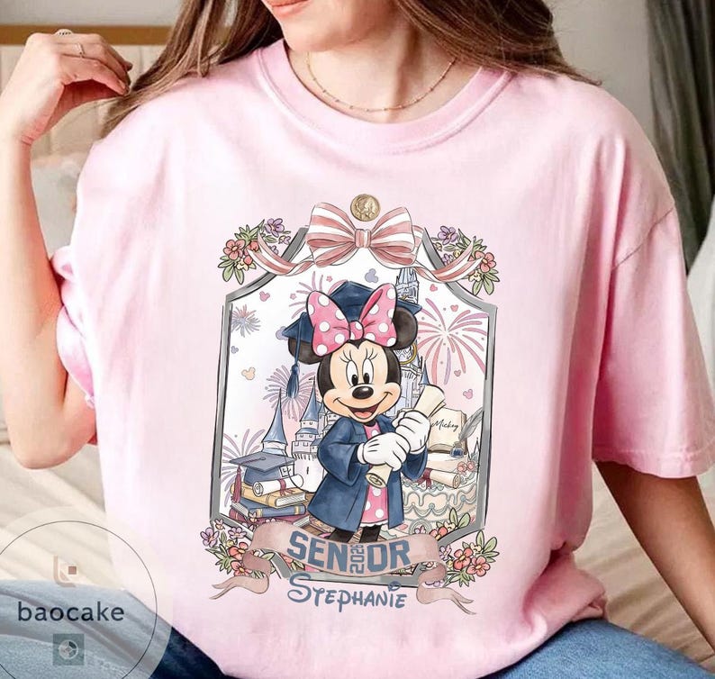 May include: Pink t-shirt featuring a Minnie Mouse graduation design. Minnie Mouse is wearing a graduation cap and gown, holding a diploma. The design includes the text "Senior Stephanie" and floral accents, with a castle and fireworks in the background.