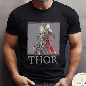 May include: Black t-shirt featuring a graphic of Thor, a superhero with blonde hair, holding a hammer. The image is framed in a gray box, with the name "THOR" printed below in a serif font.