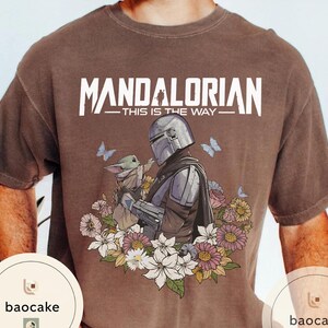 Puede incluir: Camiseta marr&oacute;n con un dise&ntilde;o de The Mandalorian y Grogu, rodeados de flores y mariposas. El texto "MANDALORIAN - THIS IS THE WAY" est&aacute; impreso en blanco en la parte superior.
