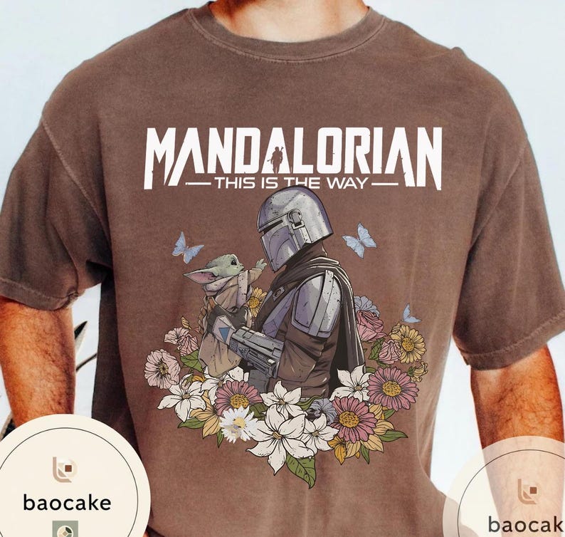 Puede incluir: Camiseta marr&oacute;n con un dise&ntilde;o de The Mandalorian y Grogu, rodeados de flores y mariposas. El texto "MANDALORIAN - THIS IS THE WAY" est&aacute; impreso en blanco en la parte superior.