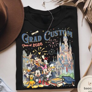 May include: Black t-shirt with a colourful graphic of Disney characters in graduation attire, with the words "Grad Custom Class of 2026". The design includes a castle, fireworks, and graduation caps.