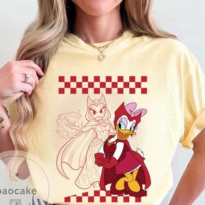 May include: Yellow t-shirt featuring a red and white checkered pattern and a graphic of two cartoon characters. One character is a duck in a red outfit with a pink bow. The other is a duck in a red and white outfit.