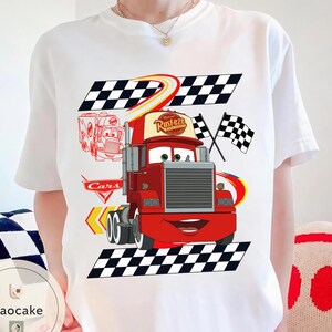 May include: White t-shirt featuring a cartoon image of the character Mack from the movie Cars. The design includes a red truck with the text "Rust-eze" on the cap, checkered flags, and the word "Cars".