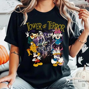 May include: Black t-shirt featuring the text "Tower of Terror" in gold lettering. The graphic includes cartoon characters like Mickey Mouse, Minnie Mouse, Donald Duck, and Goofy, with a background of a building and lightning.