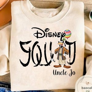 Puede incluir: Sudadera color crema con el texto "Disney Squad" en escritura negra, con un personaje de dibujos animados de Goofy sosteniendo un globo de arco&iacute;ris. Las palabras "Uncle Jo" est&aacute;n impresas debajo.