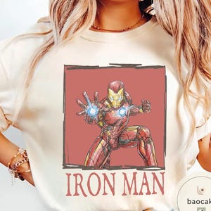 May include: Cream-colored sweatshirt featuring a red and yellow Iron Man graphic. The illustration shows Iron Man in a dynamic pose with a glowing blue energy blast. The words "IRON MAN" are printed in red below the image.