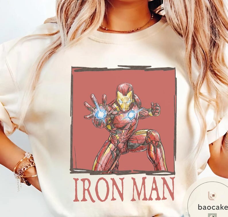May include: Cream-colored sweatshirt featuring a red and yellow Iron Man graphic. The illustration shows Iron Man in a dynamic pose with a glowing blue energy blast. The words "IRON MAN" are printed in red below the image.