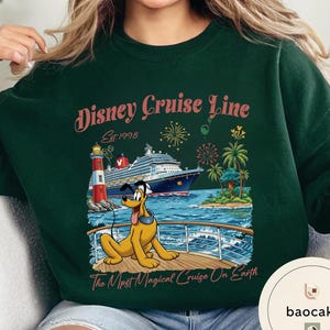 K&ouml;nnte beinhalten: Dunkelgr&uuml;nes Sweatshirt mit einem Disney Cruise Line-Motiv. Das Design zeigt ein Kreuzfahrtschiff, Pluto den Hund, einen Leuchtturm und eine tropische Landschaft. Der Text lautet "Disney Cruise Line Est. 1998" und "The Most Magical Cruise On Earth."