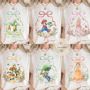 May include: A collection of six cream-colored t-shirts featuring watercolor illustrations of characters from the Super Mario video game series. Each shirt showcases a different character, including Yoshi, Mario, Princess Peach, Bowser, Luigi, and Daisy, surrounded by floral and ribbon accents.