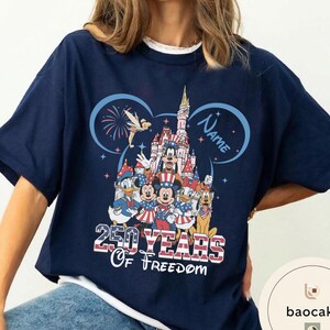 Peut inclure: T-shirt bleu marine avec un motif color&eacute; de personnages Disney en tenue patriotique devant un ch&acirc;teau. Le motif comprend le texte "250 Years Of Freedom" et un espace pour un nom. Le t-shirt a des manches courtes et un col rond.