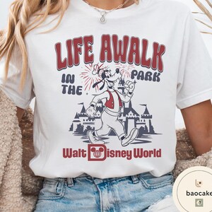 May include: White t-shirt featuring a graphic of Goofy running in front of a castle, with the text "LIFE A WALK IN THE PARK" and "Walt Disney World" in red and black.