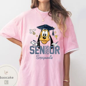 May include: Pink t-shirt with a graduation-themed design featuring a cartoon dog wearing a graduation cap. The shirt says "SENIOR 2026" and "Stephanie". The design includes stars, a trophy, and other graduation-related elements.